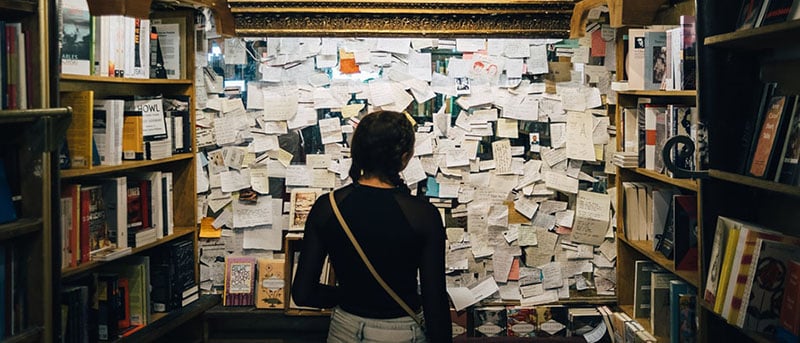 Image d'une jeune femme en train de consulter un tableau sur lequel sont épinglés des centaines de petits papiers, comme des petites annonces.
