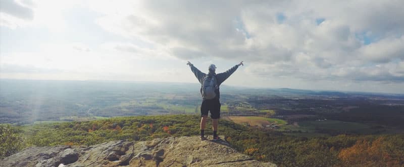 Photo d'un homme avec un sac à dos, de dos, en haut d'une colline qui lève les bras au ciel. Se retrouver premier sur google, sah quel plaisir.