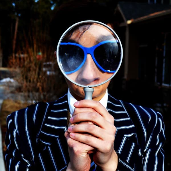 photo d'un homme en costume rayé avec des lunettes bleues, il tient une loupe devant le visage ce qui agrandit ses lunettes et son nez