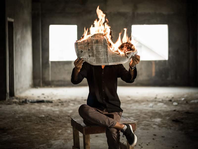 Homme assis sur une chaise dans un bâtiment désaffecté en train de lire un journal qui brûle 